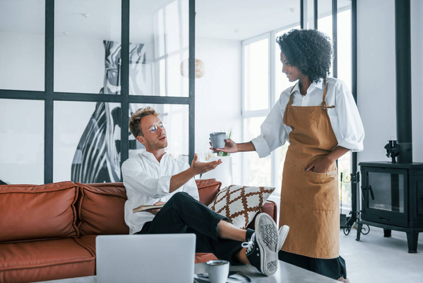 African american woman gives cup with drink. Caucasian young guy in elegant white shirt indoors at home. - Foto, afbeelding