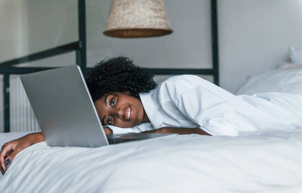 Lying down on bed with laptop. Young african american woman with curly hair indoors at home. - Φωτογραφία, εικόνα