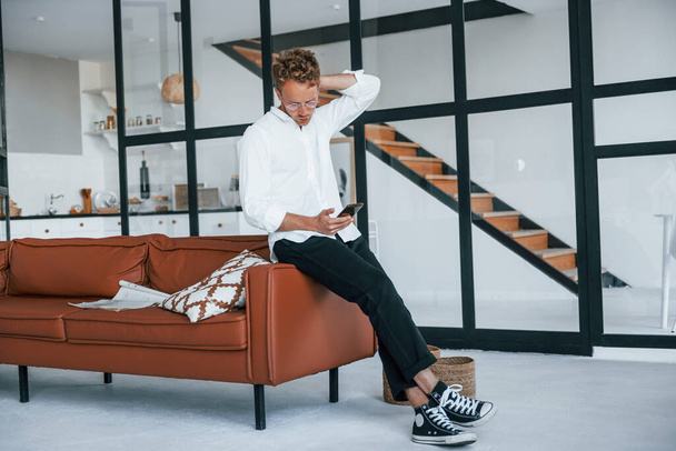 Holds phone in hand. Caucasian young guy in elegant white shirt indoors at home. - Valokuva, kuva