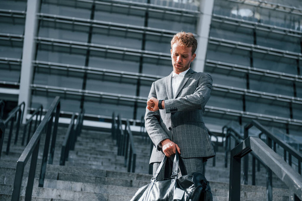 On the stairs of stadium. Young businessman in grey formal wear is outdoors in the city. - Foto, Imagen