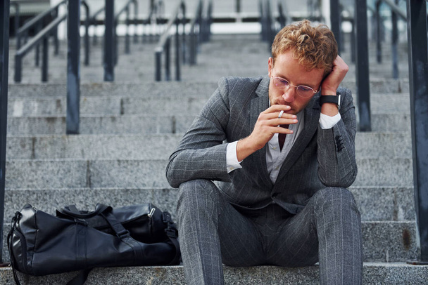 Smoking cigarette. Young businessman in grey formal wear is outdoors in the city. - Foto, imagen