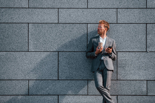 Against grey wall. Young businessman in grey formal wear is outdoors in the city. - Foto, imagen