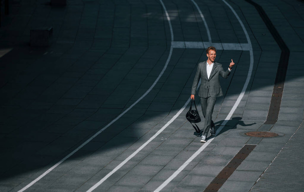 Walks on the road. Young businessman in grey formal wear is outdoors in the city. - Zdjęcie, obraz