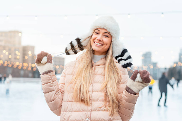 Beautiful lovely middle-aged girl blonde hair warm winter jackets knitted glove stands ice rink background Town Square. Christmas mood lifestyle Happy holiday woman walk snowy day Winter leisure - Фото, изображение