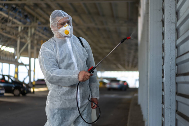 Worker wearing protective suit disinfection gear disinfect surface public place parking. Side view of disinfector safety overall and respiratory mask Covid pandemic disinfection preventive measures - Photo, image