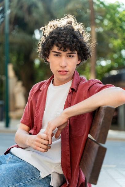 Vertical view of the handsome boy with curly hair sitting at the bench and looking attentively at the camera while relaxing at the street - Foto, afbeelding