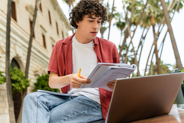 Portrait d'un étudiant masculin habillé à la mode regardant attentivement le carnet et l'ordinateur portable tout en apprenant sur un banc urbain en ville  - Photo, image