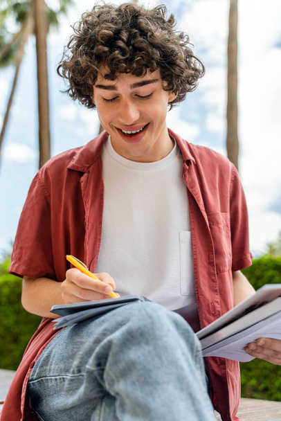 Vertical view of the curly man writing notes while sitting at the street outdoors during the great sunny day. Education concept  - 写真・画像