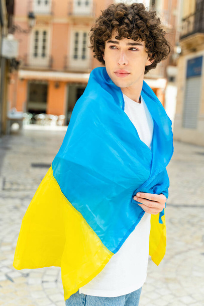 Joven hombre rizado envuelto en los colores de la bandera ucraniana en la calle, protestando por la invasión de Ucrania por Rusia  - Foto, imagen