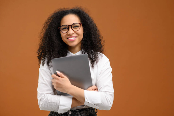 Young African American woman with afro hairstyle wearing smart casual wear and stylish eyeglasses standing isolated on brown , carrying laptop computer, smiling, female office employee - Foto, immagini