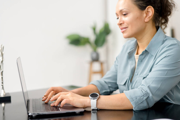 Female employee in smart casual wear looks at the camera and smiles, typing messages, answering email, front view. Confident middle-aged woman work with a laptop - Foto, immagini