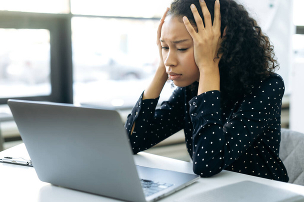 Femme afro-américaine épuisée, auditeur, avocat, économiste, assise devant un ordinateur à un bureau de travail dans un bureau moderne, tient sa tête, ne peut pas faire face à une tâche difficile, a besoin d'aide et de repos - Photo, image