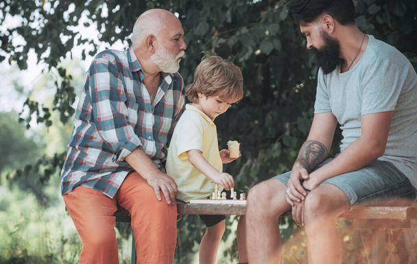 Handsome grandpa and grandson are playing chess while spending time together outdoor. Boy with father and grandfather. Father and son in the park. - Photo, Image
