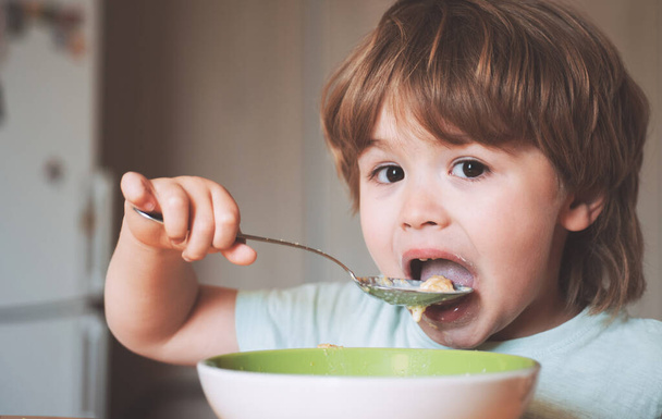 Hungriger kleiner Junge beim Essen. Fröhliches Kleinkind isst Lebensmittel selbst mit Löffel. Leckeres Kinderfrühstück. Babynahrung in der Küche. Glücklicher Junge isst gesundes Essen selbst. - Foto, Bild