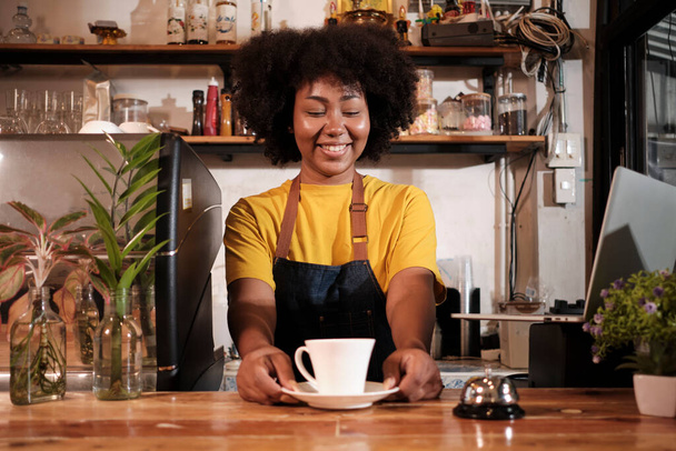 Afroamerykanka barista oferuje filiżankę kawy do klienta z wesołym uśmiechem, happy service działa w swobodnej restauracji kawiarni, młody mały przedsiębiorca start-up. - Zdjęcie, obraz