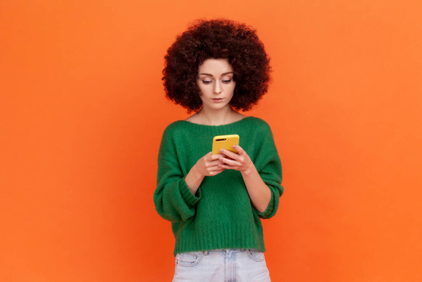 Portrait of attractive young adult woman with Afro hairstyle wearing green casual style sweater using smart phone, browsing internet, checking emails. Indoor studio shot isolated on orange background. - Foto, immagini