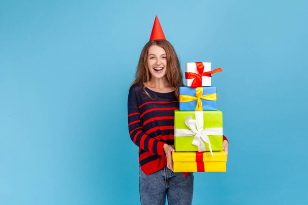 Smiling woman wearing striped casual style sweater, holding stack of presents, looking at camera with excited expression, celebrating birthday. Indoor studio shot isolated on blue background. - Photo, Image