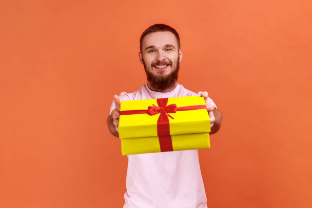 Portrait of happy bearded man giving gift box to camera with excited smile, greeting on holiday and sharing present, wearing pink T-shirt. Indoor studio shot isolated on orange background. - Fotografie, Obrázek