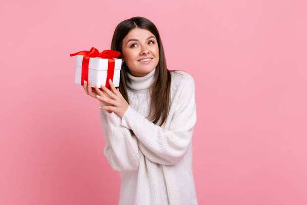 Good looking girl with dark hair holding wrapped present box, being interested what inside, smiling, wearing white casual style sweater. Indoor studio shot isolated on pink background. - Photo, Image