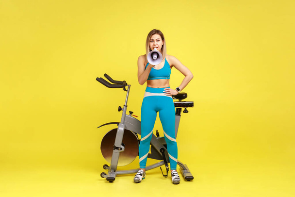 Full length portrait of sporty woman holding megaphone and making announce, inviting potential clients to fitness club, wearing blue sportswear. Indoor studio shot isolated on yellow background. - Foto, Imagen