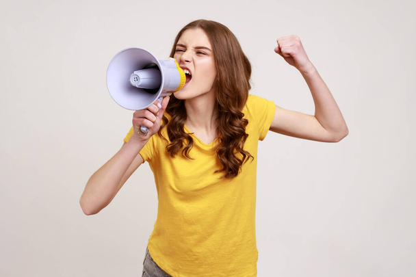 Angry nervous teenager girl with brown hair in yellow t-shirt loudly screaming at megaphone, making announce, protesting, wants to be heard. Indoor studio shot isolated on gray background. - Photo, image