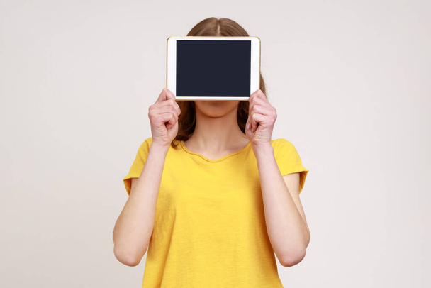 Portrét neznámé ženy ve žlutém neformálním stylu tričko zakrývající tvář s digitálním tabletem s prázdnou obrazovkou, kopírovací prostor pro reklamu. Vnitřní studio záběr izolované na šedém pozadí. - Fotografie, Obrázek