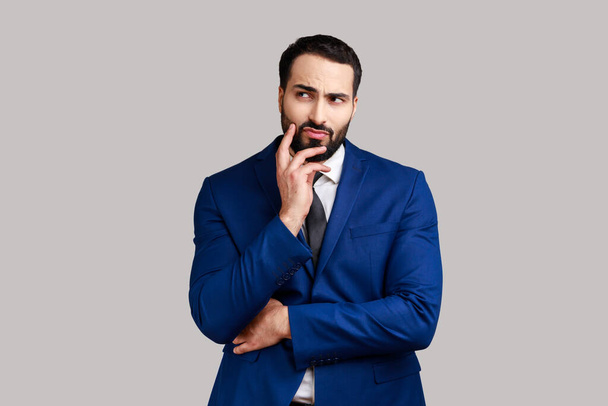 Pensive bearded businessman touching chin and thinking about important question, making hard decision, wearing official style suit. Indoor studio shot isolated on gray background. - Foto, Imagem