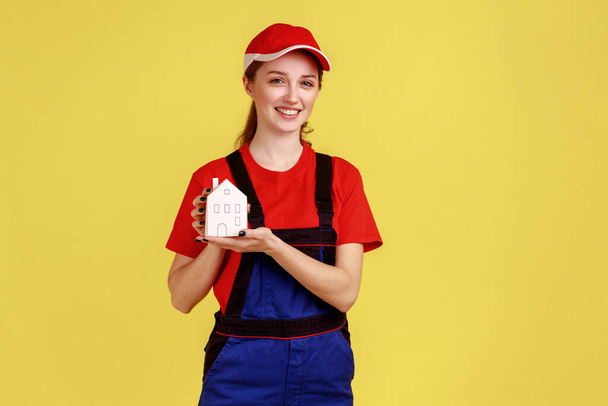 Portrait of woman mover standing holding paper house in hands, home repair services and maintenance, wearing overalls and red cap. Indoor studio shot isolated on yellow background. - Fotoğraf, Görsel