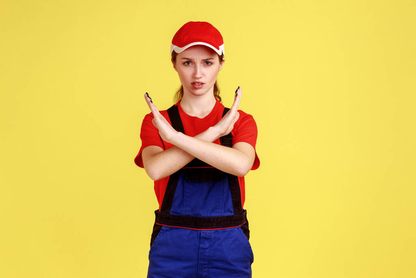 Strict bossy worker man standing and looking at camera with serious expression, showing x sign gesture, wearing overalls and red cap. Indoor studio shot isolated on yellow background. - Foto, Imagen