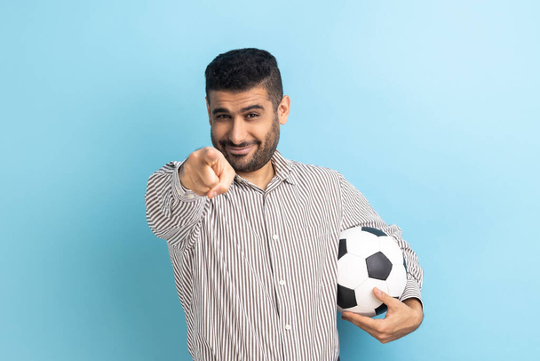 Portrait of young adult bearded businessman pointing on you, holding soccer ball, looking at camera with happy look, wearing striped shirt. Indoor studio shot isolated on blue background. - Фото, зображення