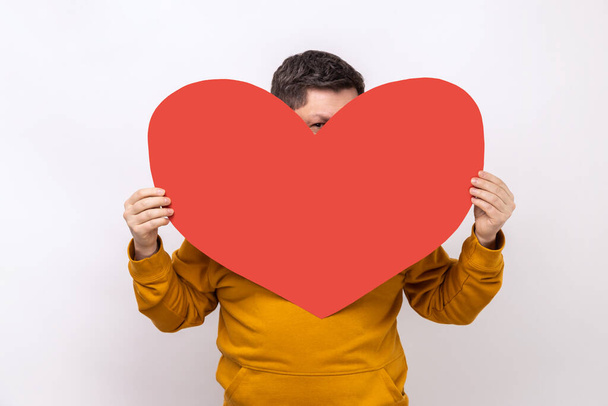 Portrait of man hiding face behind big red heart and looking at camera with curious prying eyes, feeling affection fondness, wearing urban style hoodie. Indoor studio shot isolated on white background - Foto, Bild
