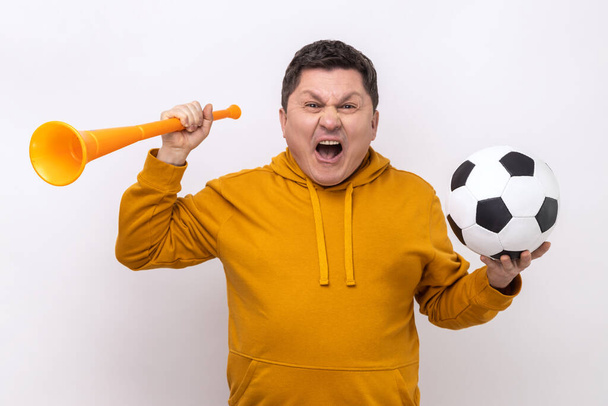 Very happy cheerful middle aged man blowing in big bullhorn holding football ball, celebrating championship beginning, wearing urban style hoodie. Indoor studio shot isolated on white background. - Foto, imagen