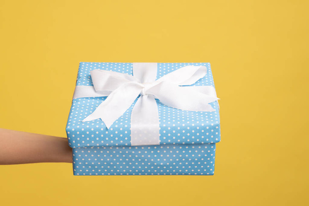 Closeup side view of woman hand holding out blue gift box with ribbon, giving present on holiday, bonuses and surprises concept. Indoor studio shot isolated on yellow background. - Fotó, kép
