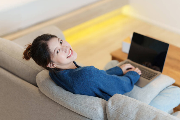 Портрет молодой азиатки, использующей социальные сети на ноутбуке. Любите делать покупки онлайн, отдыхайте на диване. - Фото, изображение