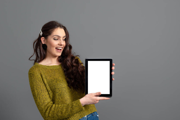 emotional young woman showing tablet computer with empty touch screen with copy space, isolated on gray background - Photo, image