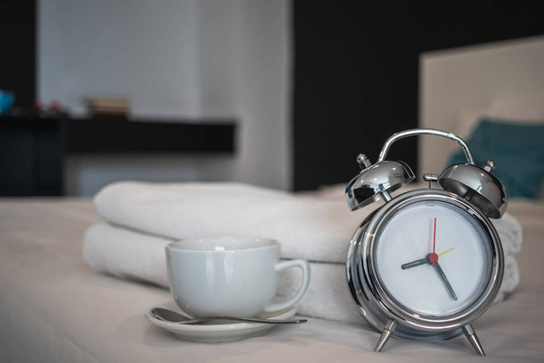 М'яке світло і розмите зображення, вінтажний годинник тривоги сидить поруч з білою керамічною чашкою кави на ліжку вранці, як чашка з кавою, підготовленою для гостей, щоб використовувати після пробудження вранці
. - Фото, зображення
