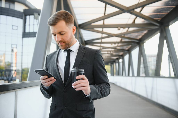 Bearded businessman in formal suit on break using mobile phone use smartphone. business man standing outside on modern urban city street background with coffee cup in downtown outdoors. copy space. - Фото, изображение