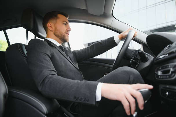 Man of style and status. Handsome young man in full suit smiling while driving a car. - 写真・画像