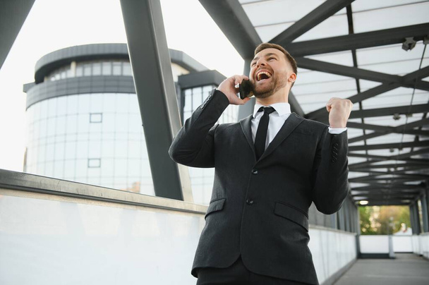 Businessman smiling and raising his fist in the air, with office building background - business success, achievement, and win concepts. - Foto, Imagen