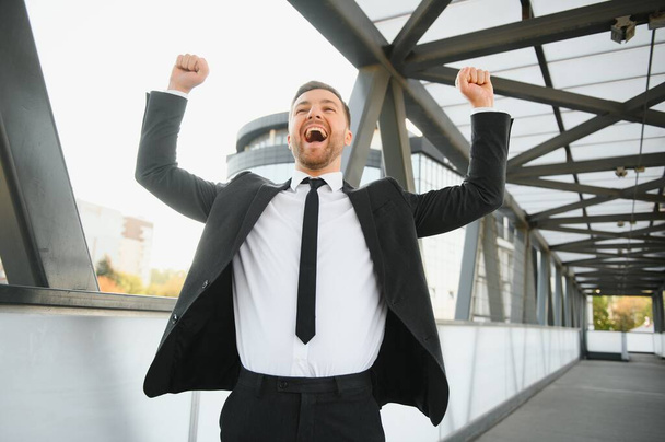 Businessman smiling and raising his fist in the air, with office building background - business success, achievement, and win concepts. - Foto, afbeelding
