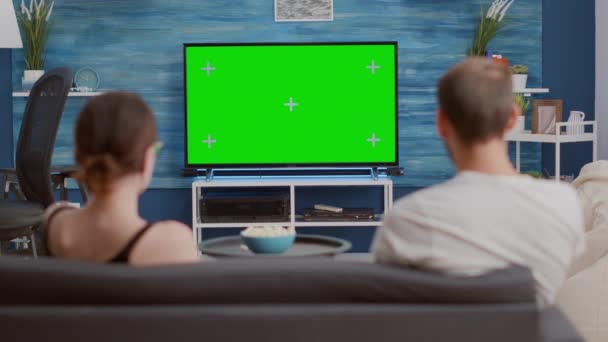 Statische Stativaufnahme eines Paares, das auf dem Sofa sitzt und auf den grünen Bildschirm im Fernseher schaut, während es sich entspannt einen Film ansieht - Filmmaterial, Video