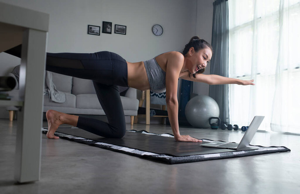 Spor yapan genç Asyalı bir kadın oturma odasında dizüstü bilgisayarın önünde yoga tahtası yapıyor, spor kıyafetleriyle spor yapan genç bir kadın evde egzersiz yapıyor, çevrimiçi fitness dersleri izliyor, oturma odasında eğitim görüyor. - Fotoğraf, Görsel
