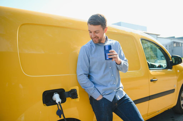 Handsome man drinking coffee while charging electric car. - 写真・画像