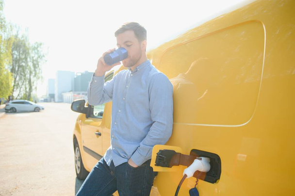 Handsome man drinking coffee while charging electric car. - 写真・画像