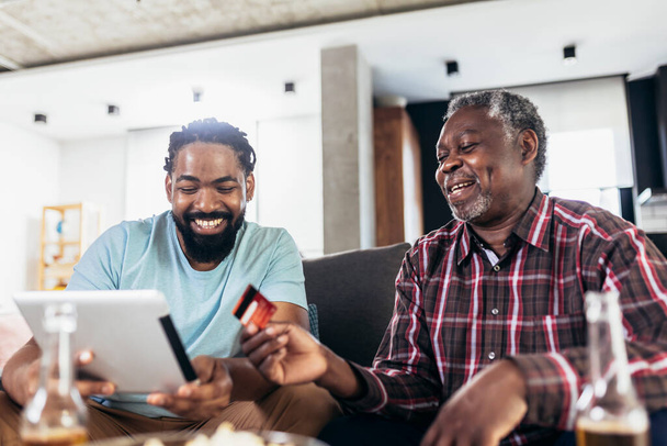 Sonriente padre e hijo usando tableta digital y tarjeta de crédito para el pago en línea en la sala de estar en casa - Foto, imagen