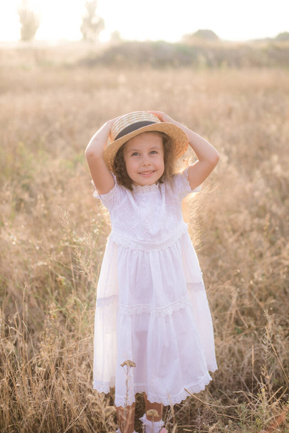 A cute little girl with long blond curly hair in a white summer dress and a straw boater hat in a field in the countryside in summer at sunset. Nature and Ecolife - Photo, image