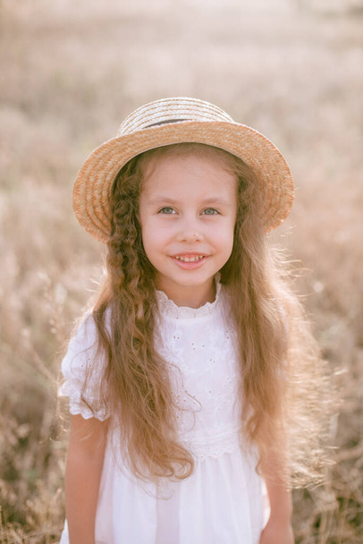 A cute little girl with long blond curly hair in a white summer dress and a straw boater hat in a field in the countryside in summer at sunset. Nature and Ecolife - Fotó, kép