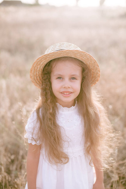 A cute little girl with long blond curly hair in a white summer dress and a straw boater hat in a field in the countryside in summer at sunset. Nature and Ecolife - Foto, afbeelding