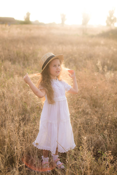 A cute little girl with long blond curly hair in a white summer dress  in a field in the countryside in summer at sunset. Nature and Ecolife - 写真・画像