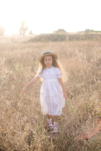 A cute little girl with long blond curly hair in a white summer dress and a straw boater hat in a field in the countryside in summer at sunset. Nature and Ecolife - Фото, изображение
